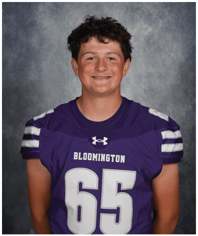 A person with short curly hair is smiling and wearing a purple football jersey with the number 65 and the word Bloomington on the front. Description generated by AI
