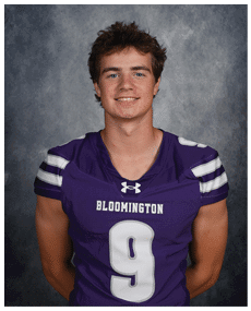 A person with short, tousled hair is smiling and wearing a purple sports jersey with white stripes on the sleeves against a gray mottled background. Description generated by AI