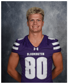A person with short blond hair is smiling and wearing a purple sports jersey with the number 80 and the word Bloomington on the front. Description generated by AI