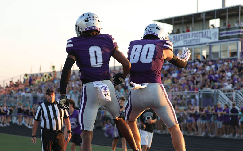 Two football players in purple and gray uniforms celebrate on the field with a referee and a crowd in the background during a game. Description generated by AI