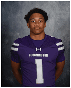 A person with short curly hair wearing a purple Bloomington football jersey with the number one stands against a gray textured background. Description generated by AI