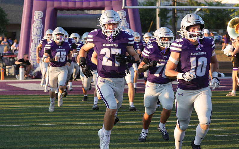 People wearing purple and white football uniforms are running onto a field from an inflatable tunnel with the word “Panthers" visible on it. Description generated by AI