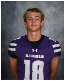 Person with short, light-colored hair wearing a purple Bloomington football jersey with the number 18 stands against a gray textured background. Description generated by AI