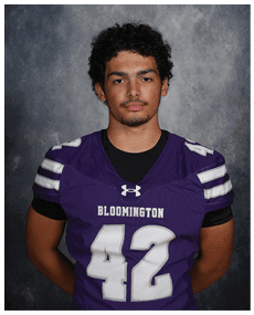 A person with curly hair wearing a purple Bloomington sports jersey with the number 42 stands against a gray textured background with hands behind their back. Description generated by AI