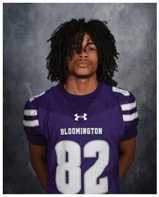 A person with medium-length curly hair is wearing a purple sports jersey with the number 82 and the word Bloomington on the front, standing against a dark textured background. Description generated by AI
