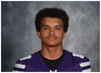 Person with short curly hair wearing a purple sports jersey with white stripes posing against a gray textured background. Description generated by AI