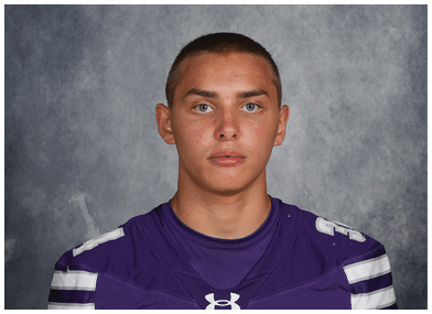 A person with short hair wearing a purple sports jersey with white stripes is looking directly at the camera against a gray textured background. Description generated by AI