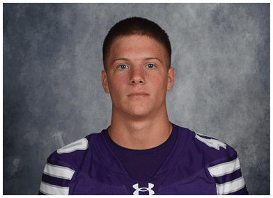 A person with short hair wearing a purple sports jersey is looking directly at the camera against a gray textured background. Description generated by AI