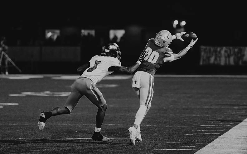 Two people in football uniforms are on a field with one person reaching out to catch a football while the other tries to defend. Description generated by AI