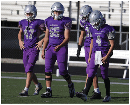 Four people wearing purple football uniforms and silver helmets stand on a field with a fence and bleachers in the background. Description generated by AI
