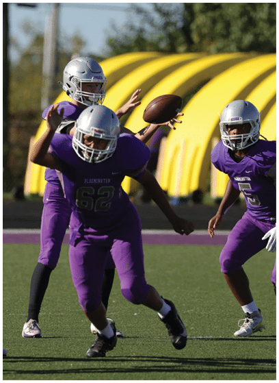 Three people in purple football uniforms and silver helmets are playing on a field with one person throwing a football. Description generated by AI