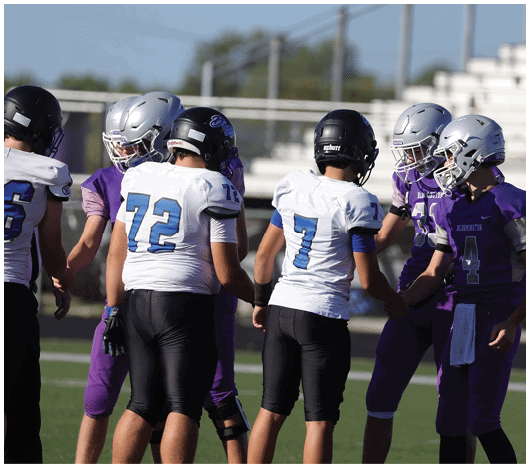 People wearing football helmets and uniforms are standing on a field, with some shaking hands. Description generated by AI
