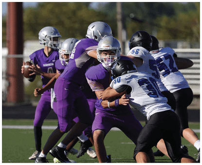 Several people wearing football helmets and uniforms are engaged in a game or practice outdoors with trees and a utility pole in the background. Description generated by AI