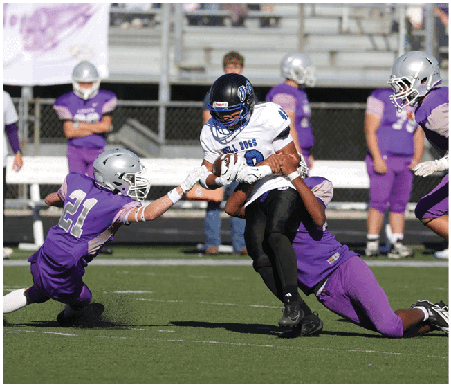 A person in a white and black football uniform is holding the ball while being tackled by two people in purple football uniforms on a green field. Description generated by AI
