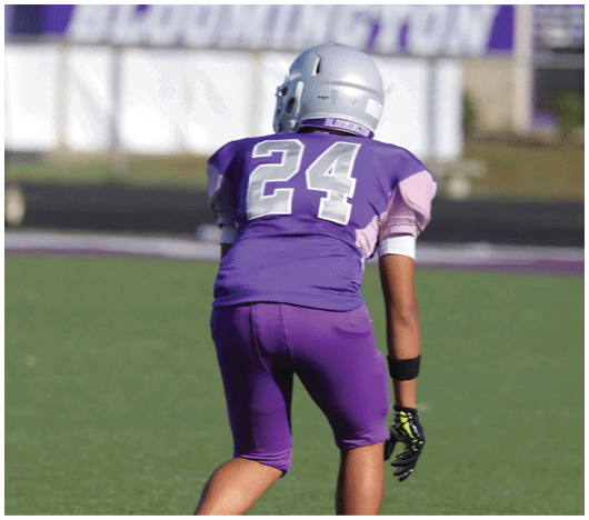 A person wearing a purple football uniform with the number 24 and a silver helmet is standing on a football field with a blurred “BLOOMINGTON" sign in the background. Description generated by AI