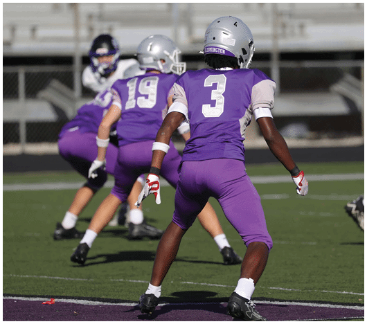 People wearing purple sports uniforms are positioned on a football field, appearing to prepare for a play. Description generated by AI