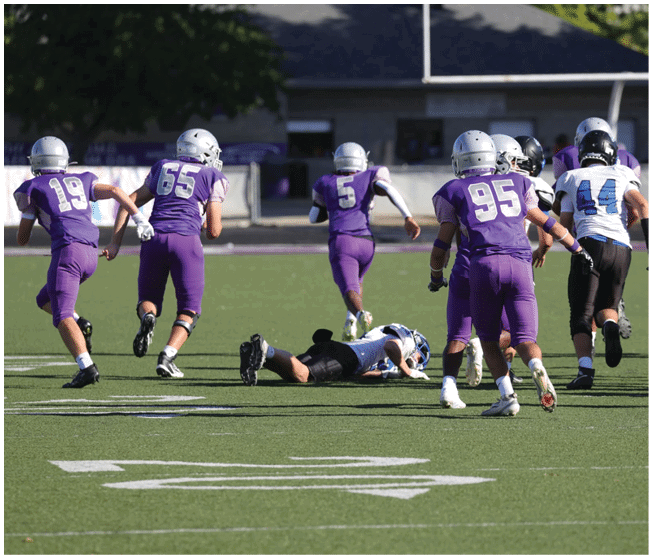 People wearing helmets and sports uniforms are playing football on a field with one person lying on the ground and others running. Description generated by AI