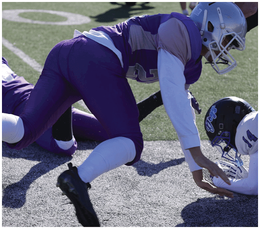 Two people in purple and white football uniforms are on a field, with one person lying on the ground and the other leaning forward. Description generated by AI