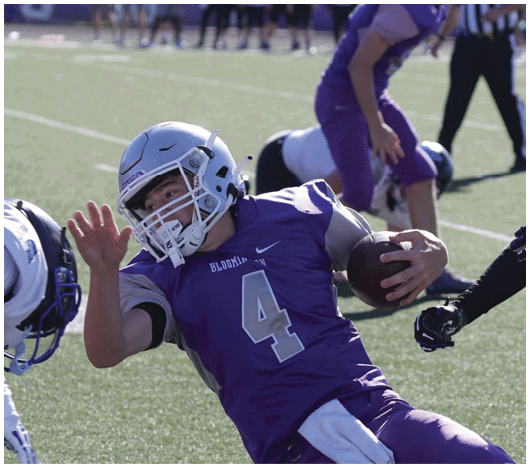 A person wearing a purple football uniform with the number 4 is holding a football and falling backward while another person in a white uniform leans forward on a football field. Description generated by AI
