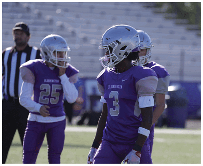 People wearing purple sports uniforms and cleats are standing on a grassy field, appearing to prepare for or participate in an athletic activity. Description generated by AI