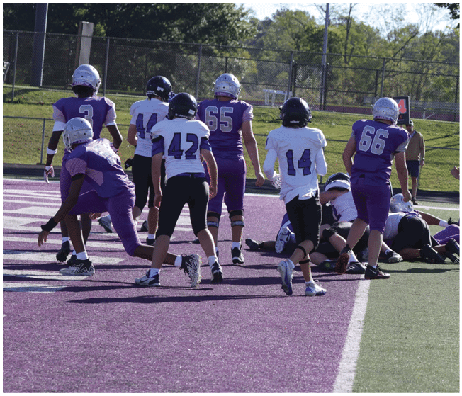 People wearing football uniforms are gathered on a field with a purple end zone and green turf, some standing and others on the ground during a game. Description generated by AI