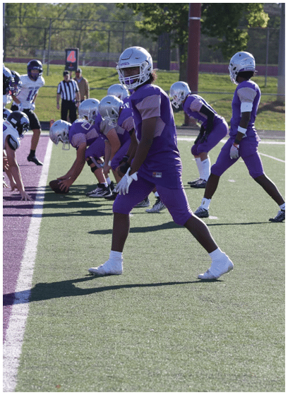 A group of people in purple and white football uniforms are lined up on a field preparing for a play during a game. Description generated by AI
