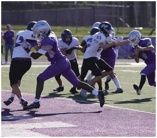 People wearing football uniforms are actively engaged in a play on a field with one player in purple tackling a player in white. Description generated by AI