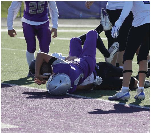 People in football uniforms are engaged in a play on a sunlit field with one person lying on the ground holding the ball and others standing nearby. Description generated by AI