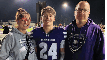 Three people smiling and posing together at a nighttime outdoor sports event, with one person wearing a football uniform and the others wearing hoodies. Description generated by AI