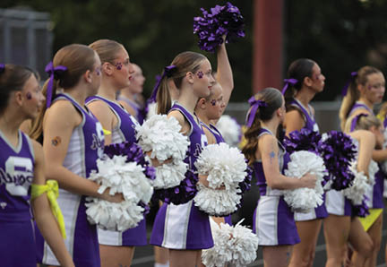 A line of people dressed in purple and white cheerleading uniforms hold white and purple pom-poms while standing outdoors. Description generated by AI