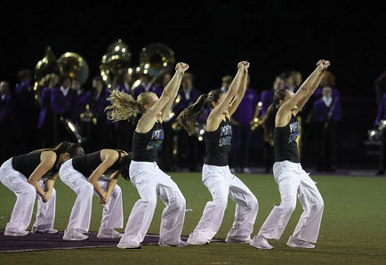 A group of people wearing black tank tops and white pants are performing a synchronized dance or cheer routine on a field with a band in purple uniforms playing instruments in the background. Description generated by AI