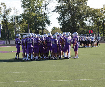 A group of people in purple football uniforms huddle together on a field while another group in white uniforms stands in the background near a scoreboard. Description generated by AI