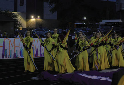 People in yellow costumes holding flags are standing in formation on a track field at night with a colorful banner in the background. Description generated by AI