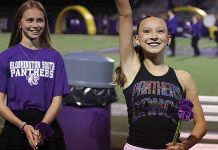 Two smiling people stand on a sports field holding purple flowers, one wearing a black tank top with colorful text and the other wearing a purple shirt with white text. Description generated by AI