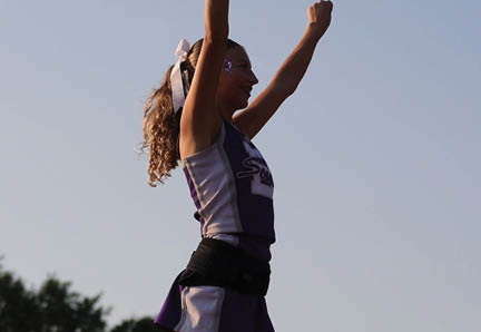 A person with curly hair tied with a ribbon is wearing a cheerleading outfit and raising both arms in the air against a clear sky. Description generated by AI