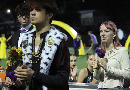 A person wearing a ceremonial robe with a fur collar and medallions stands in the foreground while two other people hold flowers in an outdoor nighttime setting. Description generated by AI