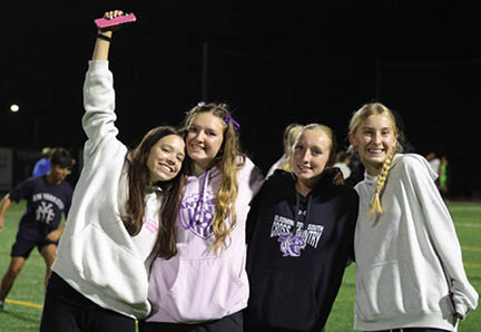 Four people wearing casual hoodies stand closely together on a sports field at night, smiling with one person raising an arm holding a pink object. Description generated by AI