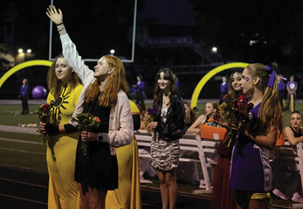 People dressed in colorful outfits stand on a sports field at night holding flowers, with one person raising a hand. Description generated by AI
