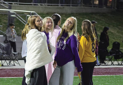 A group of people stand on a sports field at night with some wrapped in blankets while others watch from bleachers in the background. Description generated by AI
