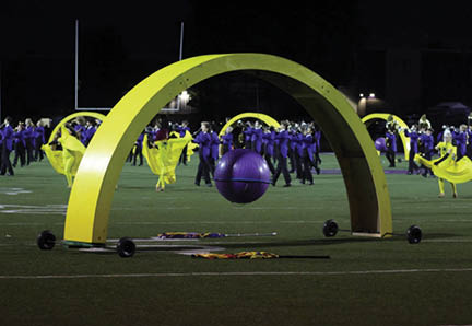 People in colorful uniforms perform a marching band routine on a field with a large yellow arch and a suspended purple ball in the foreground. Description generated by AI