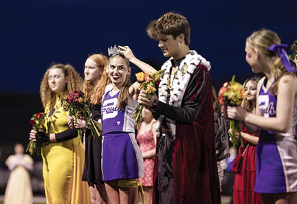A group of people dressed in colorful outfits stand in a line outdoors at night, with one person placing a crown on another who is smiling and holding flowers. Description generated by AI
