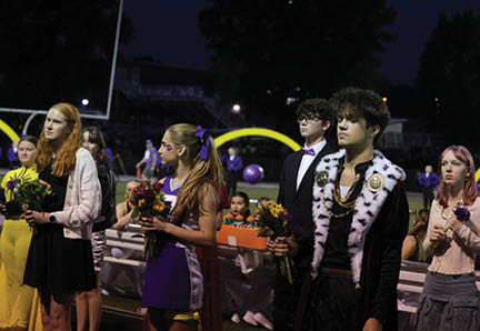 People dressed in formal and cheerleading attire stand on a sports field at night holding flowers, with some seated in the background. Description generated by AI