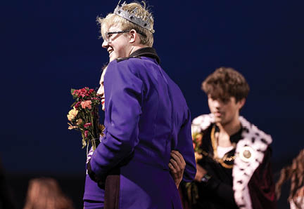 A person wearing a crown and a purple jacket holds a bouquet of flowers while smiling and embracing another person, with others in the background dressed in formal or costume attire. Description generated by AI