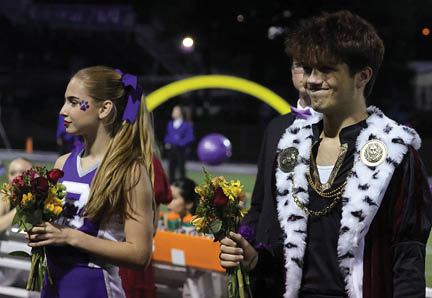 Two people dressed in a cheerleader outfit and a royal costume hold bouquets of flowers while standing outdoors at night. Description generated by AI