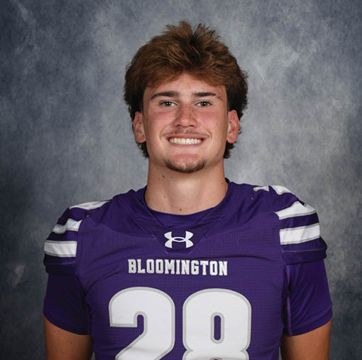 A person with short tousled hair is smiling and wearing a purple sports jersey with the number 28 and the word Bloomington on the front against a gray mottled background. Description generated by AI