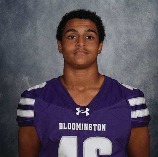 A person wearing a purple Bloomington football jersey with white stripes on the sleeves is standing against a gray textured background. Description generated by AI