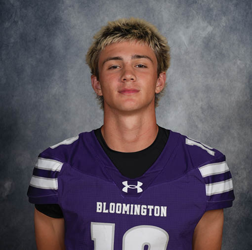 A person with short, light-colored hair is wearing a purple sports jersey with the word Bloomington and the number 18 on it, standing against a gray textured background. Description generated by AI