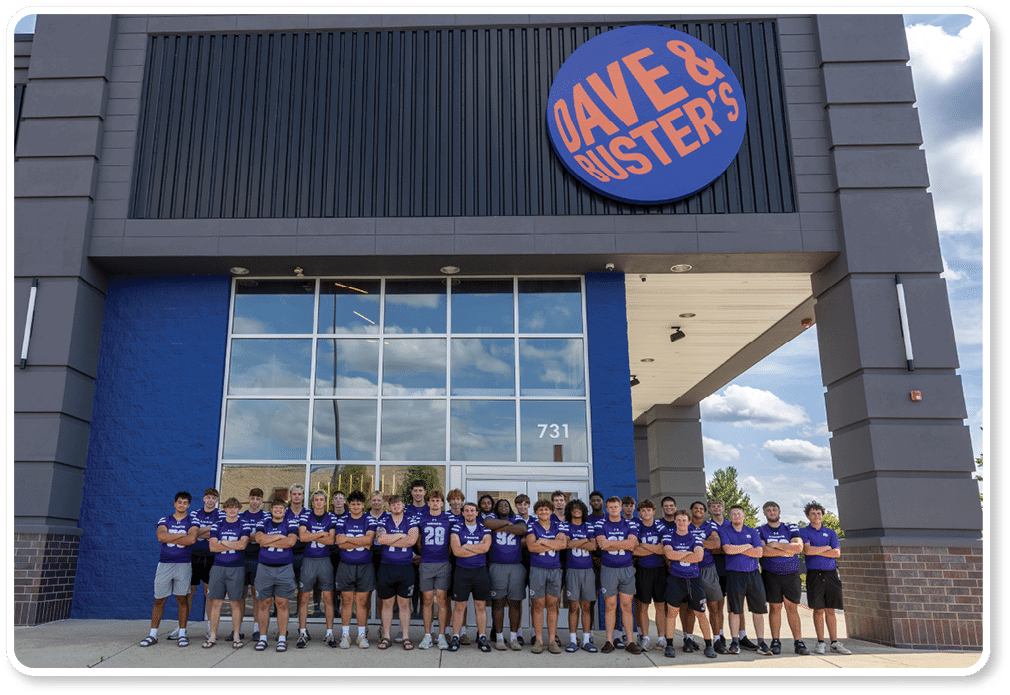 A group of people wearing matching purple shirts and shorts stand side by side in front of a blue wall with brick pillars. Description generated by AI