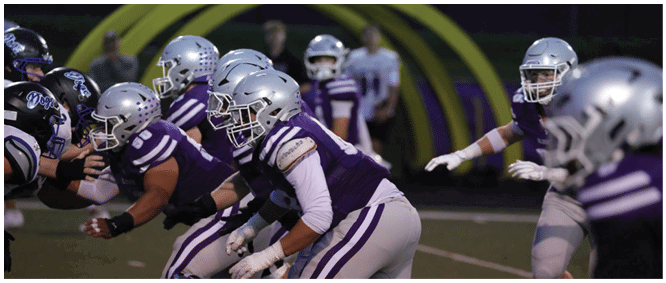 People in football uniforms and helmets are lined up on a field preparing for a play during a game. Description generated by AI