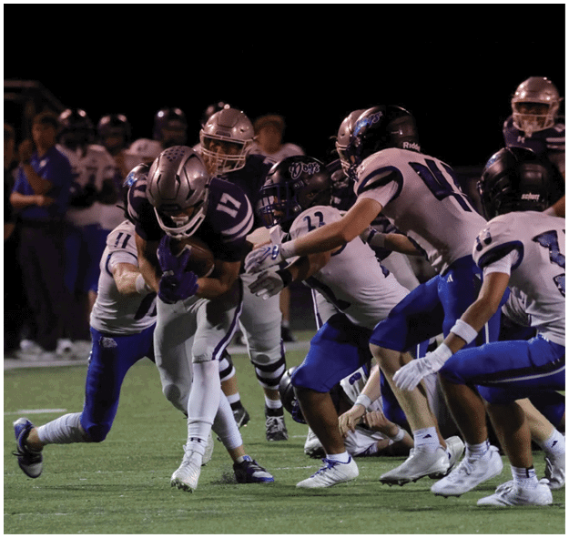 A group of people wearing football uniforms are engaged in a nighttime game with one person holding the ball while others attempt to tackle or block. Description generated by AI